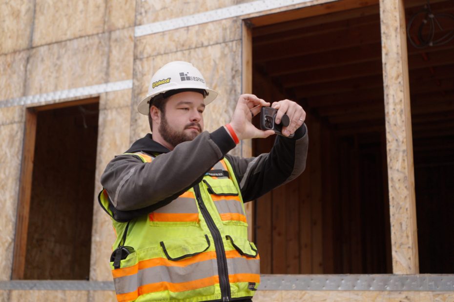 Pacific student Colin Schneider on a construction site, wearing protective gear, takes a photo on his phone.